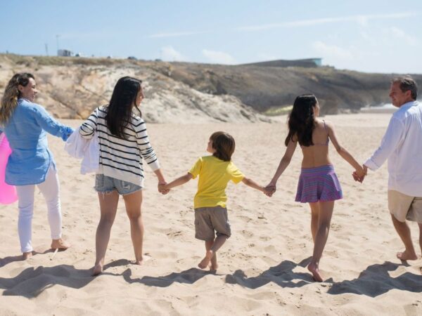 Une famille de cinq personnes vue de dos, main dans la main, qui marche sur une plage de sable avec une bouée rose et un panier