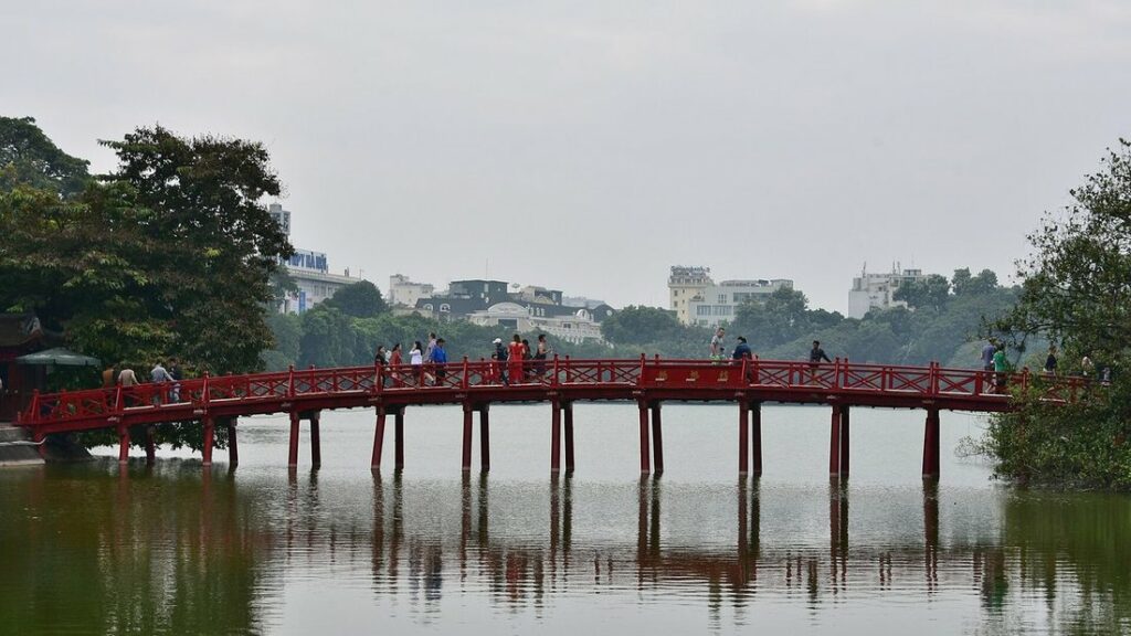 Pont rouge de The Huc sur le lac Hoan Kiem, avec arbres et immeubles de Hanoï à l'arrière-plan