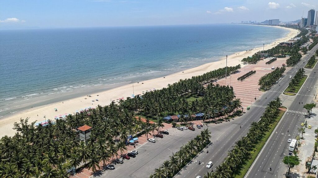 Longue plage de sable vue d'en haut, bordée d'une avenue et de palmiers, avec la mer à gauche