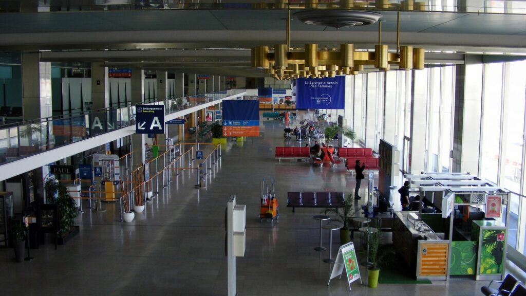 Grand hall de départ d'un aéroport presque vide, files de barrières sans voyageurs, quelques personnes assises sur des banquettes rouges