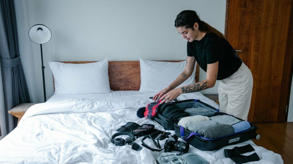 Femme penchée sur une valise ouverte posée sur un lit, avec appareil photo, écouteurs et lunettes de soleil à côté