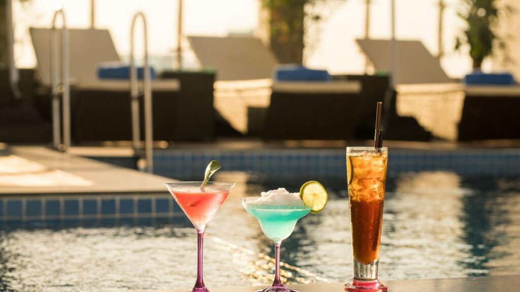 Trois boissons colorées posées au bord d'une piscine, avec des transats flous en arrière-plan