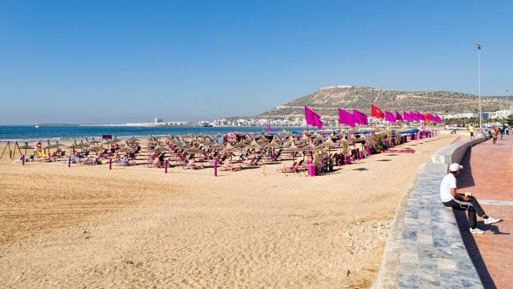 Plage d'Agadir avec parasols de paille, fanions roses et la colline qui domine la baie au fond