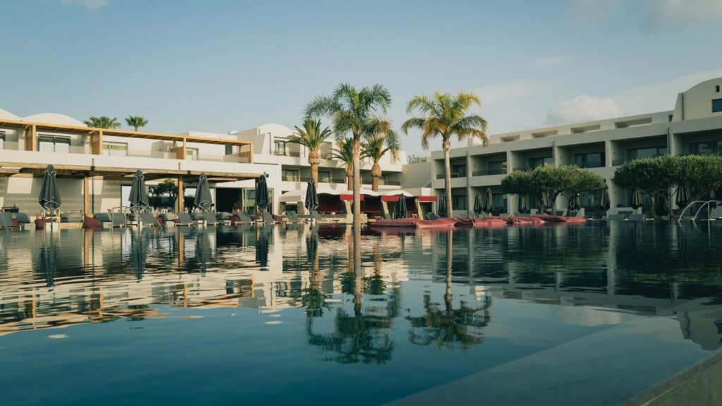 Bassin calme d'un hôtel-club reflétant des bâtiments blancs, des palmiers et des transats aux parasols fermés