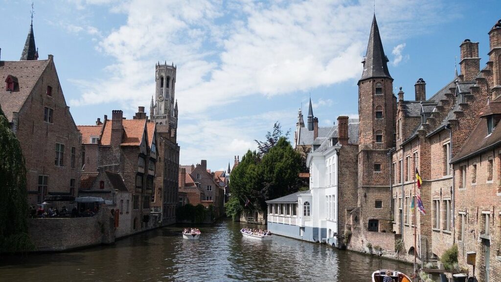 Canal de Bruges bordé de maisons de brique, avec le beffroi au loin et des bateaux de promenade sur l'eau