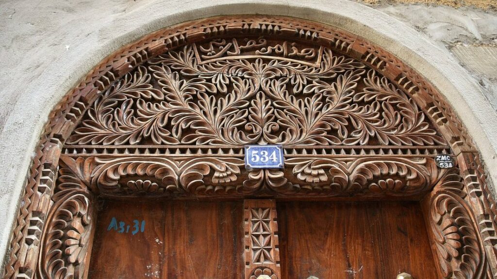 Arche de bois sculptée de motifs de feuillages au-dessus d'une porte, avec une plaque de numéro bleue, dans un mur de pierre claire