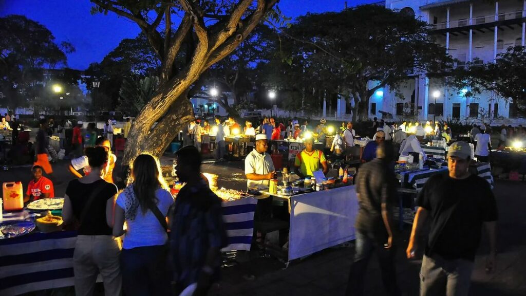 Étals éclairés et passants dans un jardin la nuit, avec de grands arbres et un bâtiment blanc au fond
