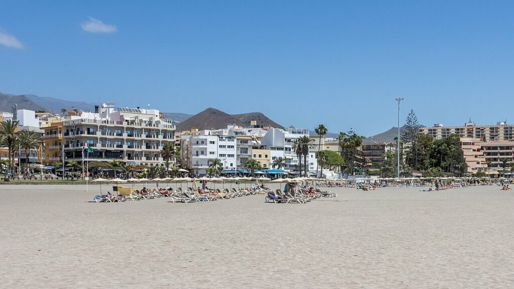 Large plage de sable clair avec transats et parasols devant des immeubles blancs et une colline sèche, à Los Cristianos