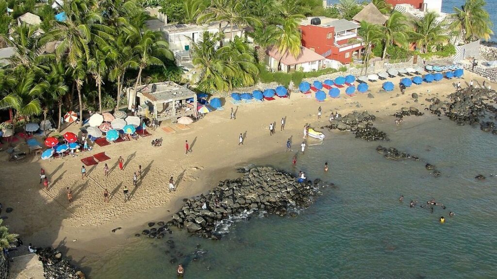 Vue en hauteur sur la plage de Ngor à Dakar, avec parasols bleus, rochers sombres, palmiers et baigneurs