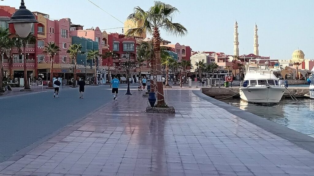 Promenade dallée de la marina de Hurghada avec palmiers, façades colorées, un yacht amarré et deux minarets au loin