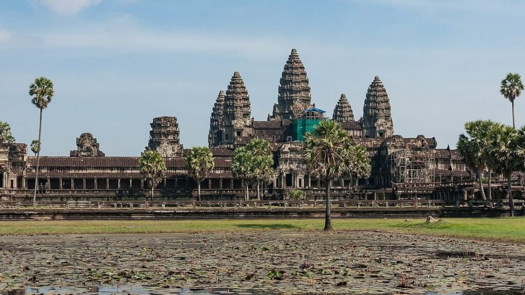 Les tours en pierre d'Angkor Wat derrière un étang couvert de nénuphars et quelques palmiers, sous un ciel bleu