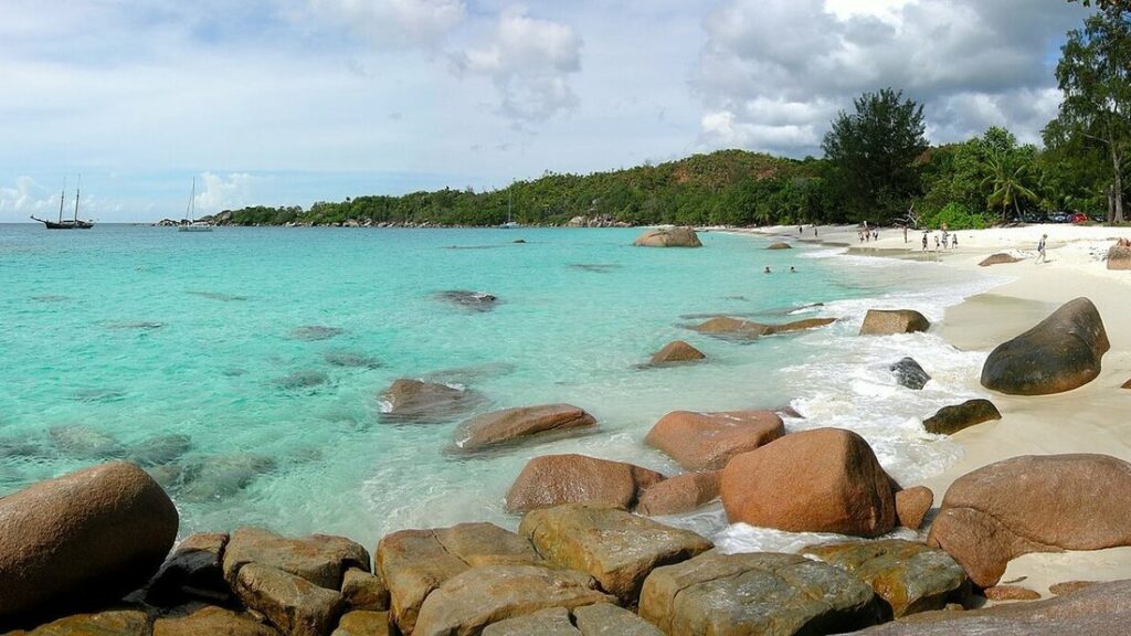 Plage de sable clair et eau turquoise bordées de gros blocs de granit, avec deux voiliers au large, aux Seychelles