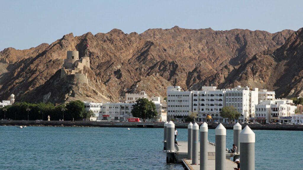 Front de mer de Mutrah à Mascate, avec des façades blanches, un petit fort sur la crête, des montagnes brunes et un ponton au premier plan au-dessus d'une eau bleu-vert
