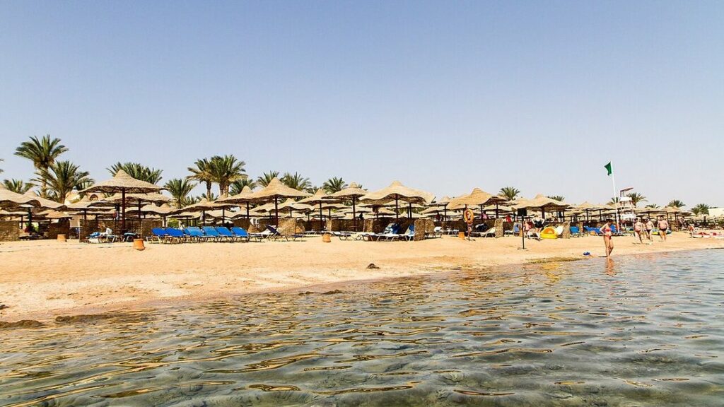 Plage de sable clair bordée de parasols de paille et de palmiers, eau peu profonde au premier plan, à Hurghada