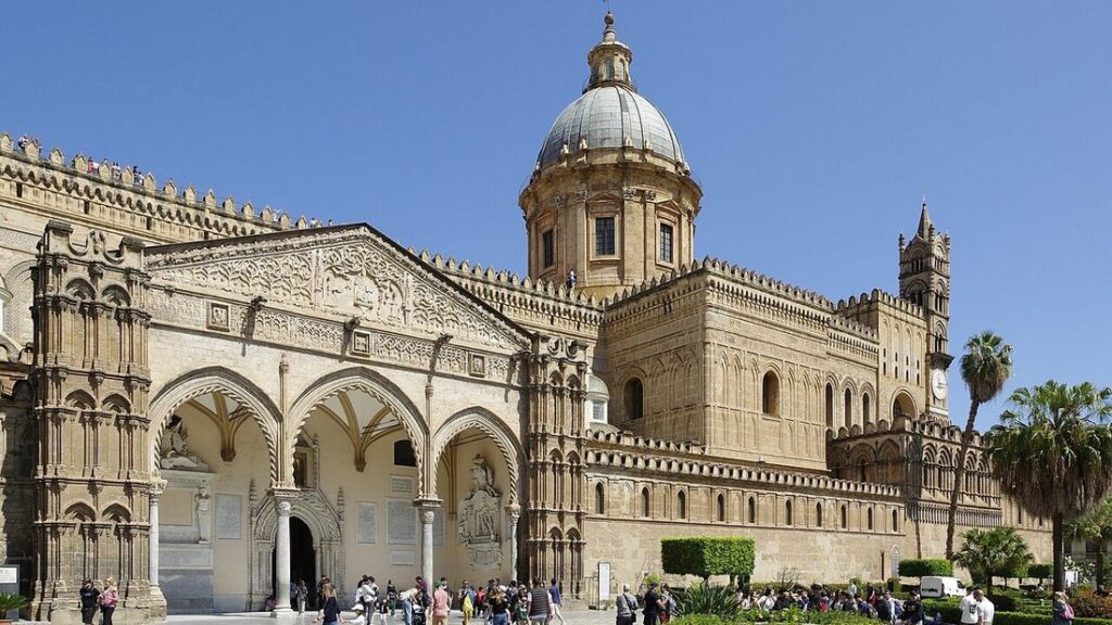 Façade de pierre dorée de la cathédrale de Palerme, avec porche à arcs gothiques, coupole et palmiers