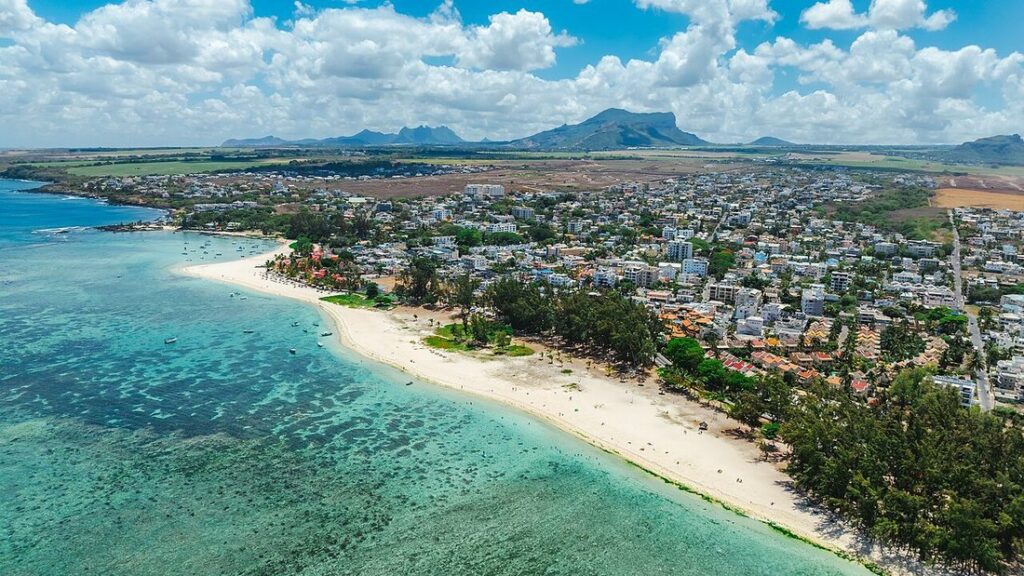 Vue aérienne d'une plage de sable blanc, d'un lagon turquoise et d'un village, montagnes au loin, à Flic-en-Flac