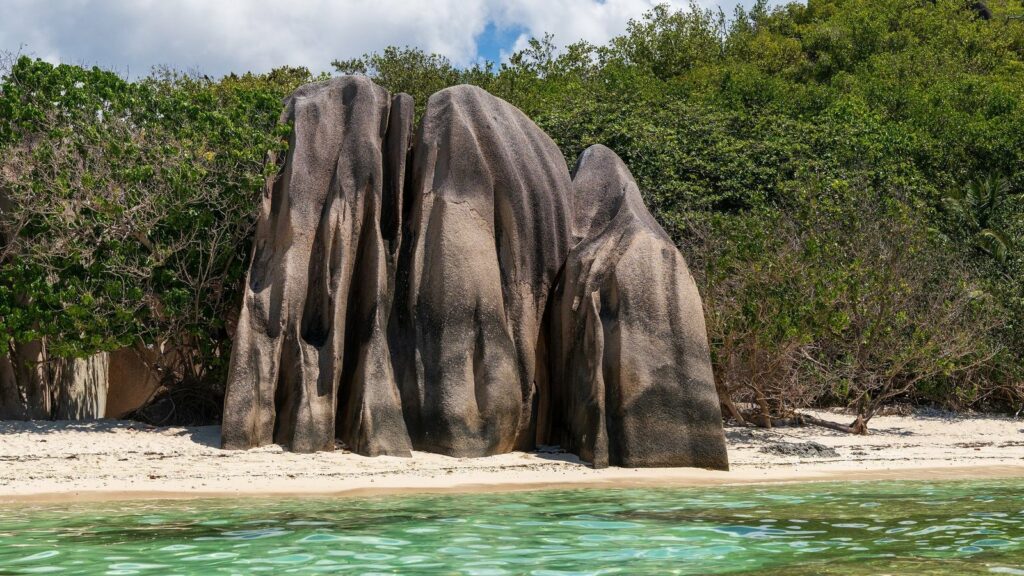 Trois rochers de granite arrondis sur une plage de sable clair bordée de végétation, avec une eau vert clair au premier plan