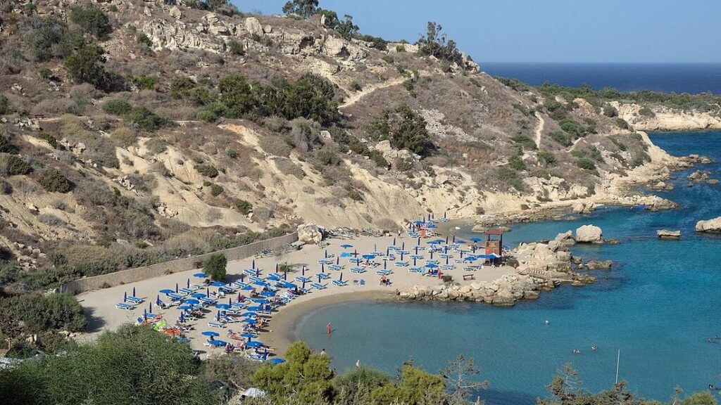 Petite plage de sable avec parasols bleus au pied d'une colline rocheuse, devant une eau turquoise