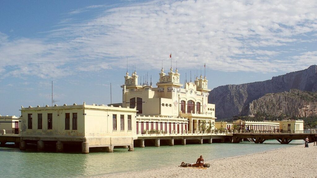 Pavillon balnéaire ancien sur pilotis au bord d'une plage de sable, avec une falaise rocheuse à l'arrière-plan
