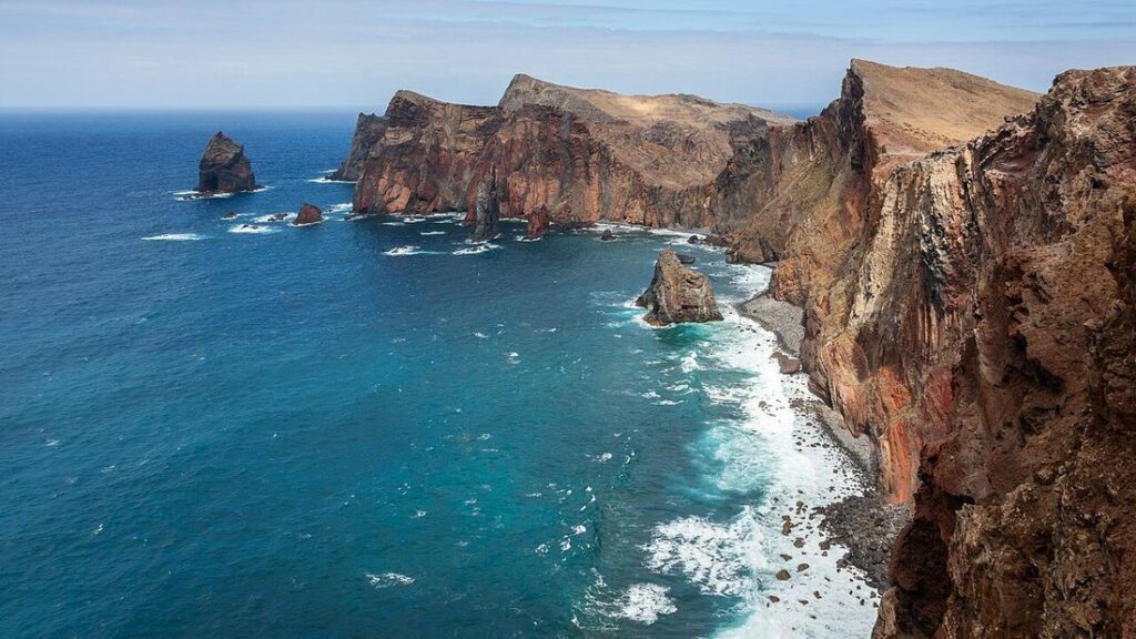 Falaises rouges et ocre plongeant dans une mer bleu profond, avec des îlots rocheux et de l'écume au pied
