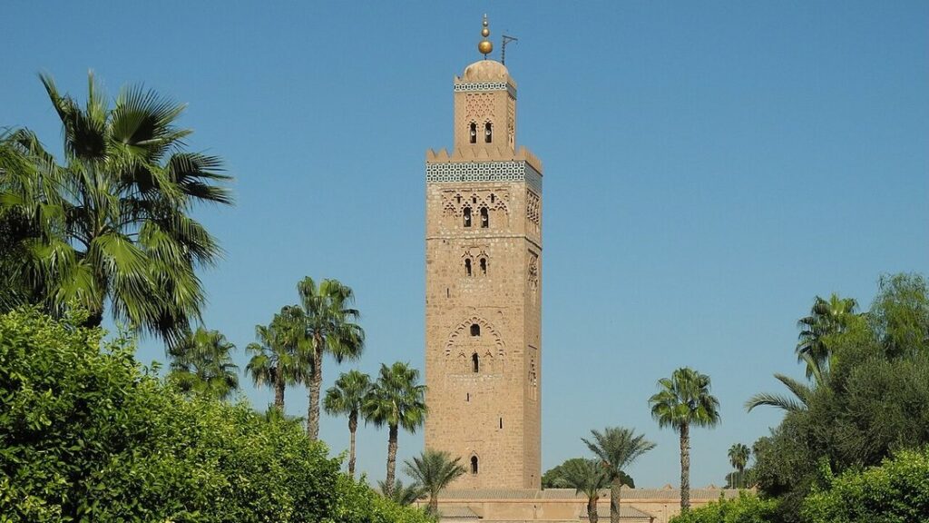Haut minaret en pierre ocre sous un ciel bleu, entouré de palmiers et de haies vertes