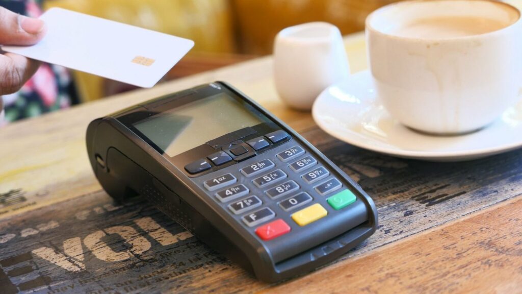 Une main tend une carte bancaire blanche au-dessus d'un terminal de paiement posé sur une table en bois, à côté d'une tasse