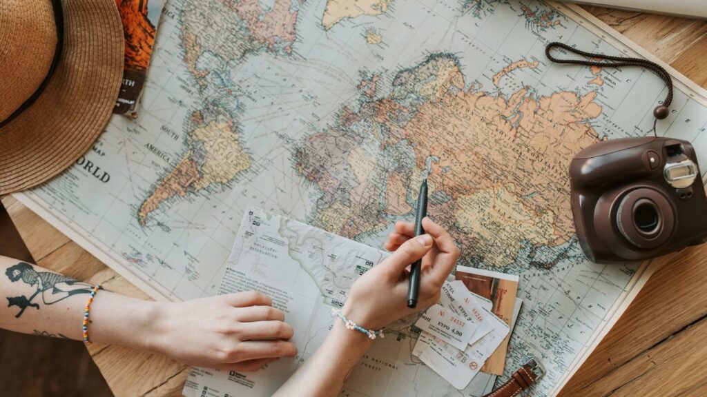 Mains au-dessus d'une carte du monde dépliée sur une table en bois, avec un stylo, des tickets, un chapeau de paille et un appareil photo