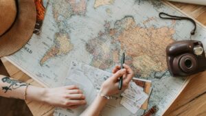 Mains au-dessus d'une carte du monde dépliée sur une table en bois, avec un stylo, des tickets, un chapeau de paille et un appareil photo