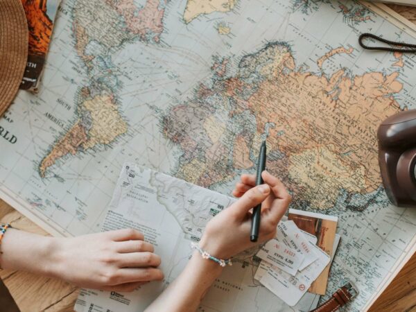 Mains au-dessus d'une carte du monde dépliée sur une table en bois, avec un stylo, des tickets, un chapeau de paille et un appareil photo