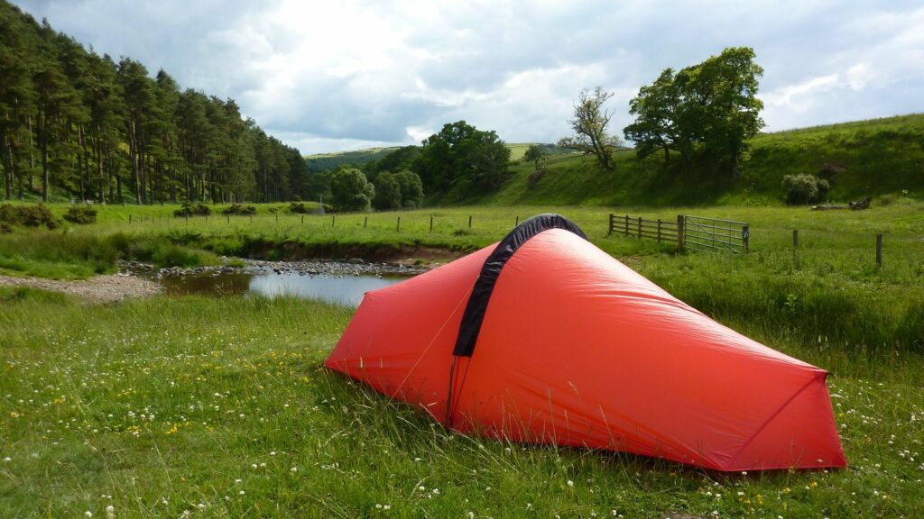 Petite tente rouge montée sur l'herbe au bord d'une rivière, dans une vallée verte bordée de pins