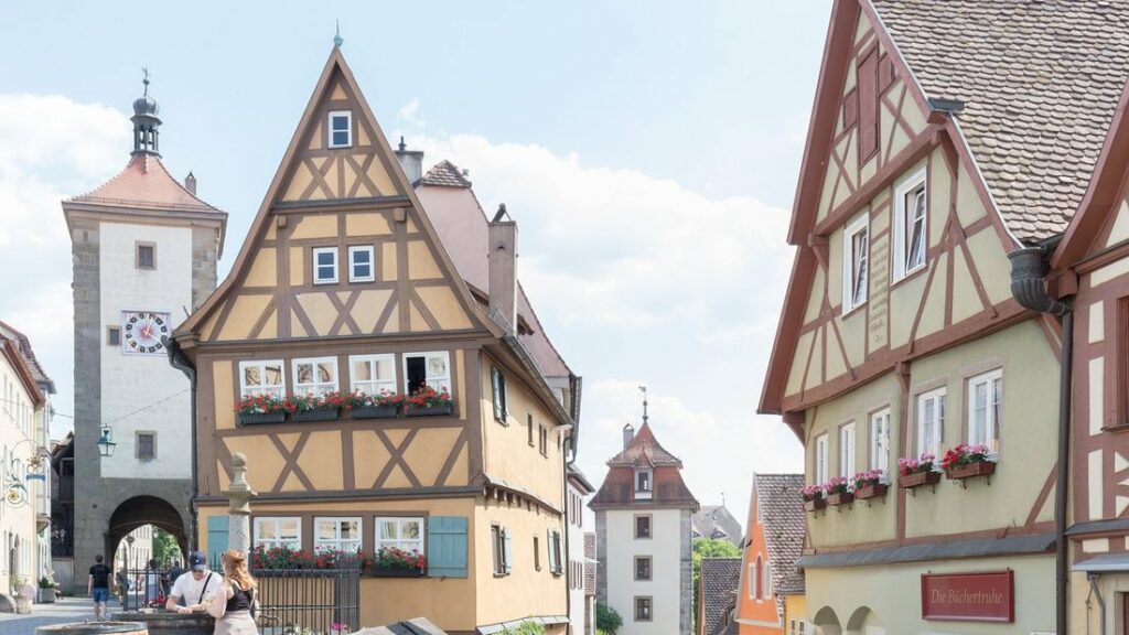 Maison à colombages jaune ornée de jardinières fleuries, entre deux tours-portes et une ruelle pavée, à Rothenburg ob der Tauber