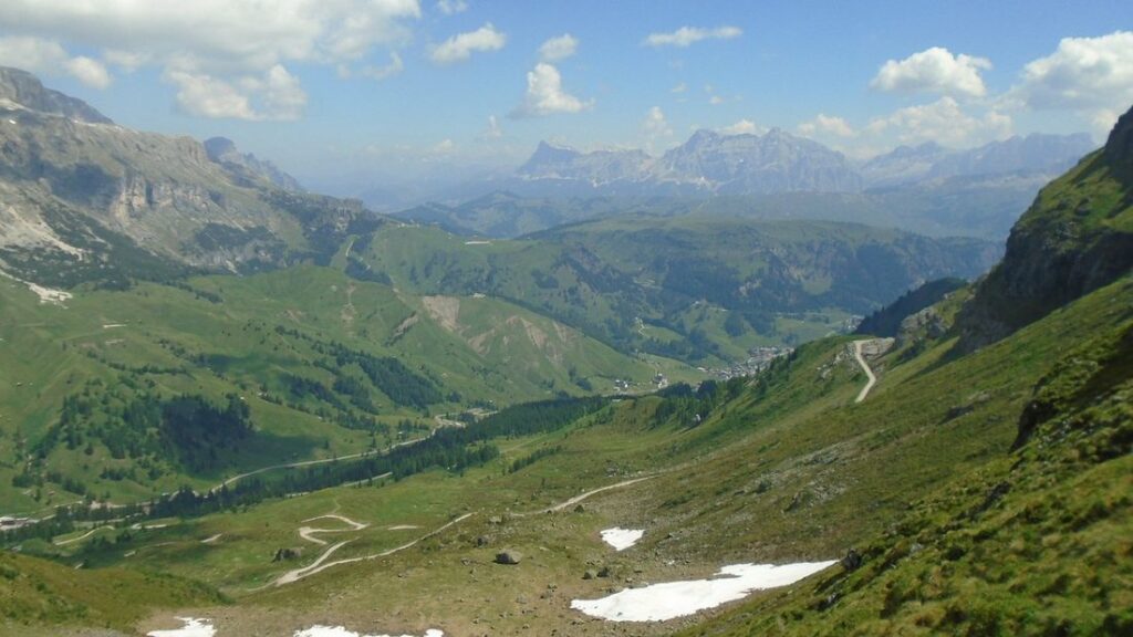 Vallée herbeuse des Dolomites vue depuis un col, avec des plaques de neige au premier plan et des sommets pâles à l'horizon