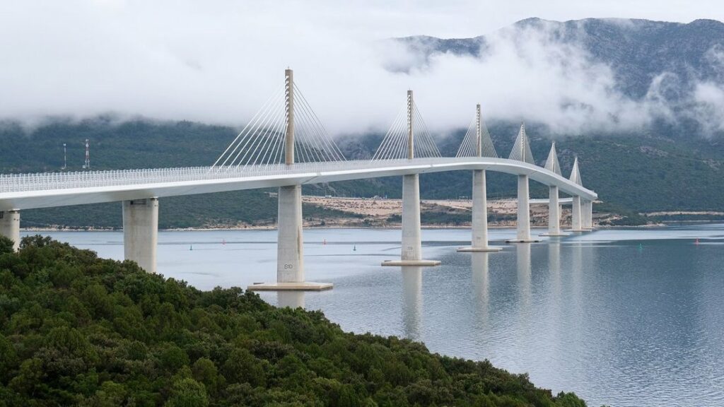 Pont haubané blanc à pylônes multiples traversant un bras de mer, avec des montagnes et des nuages bas derrière
