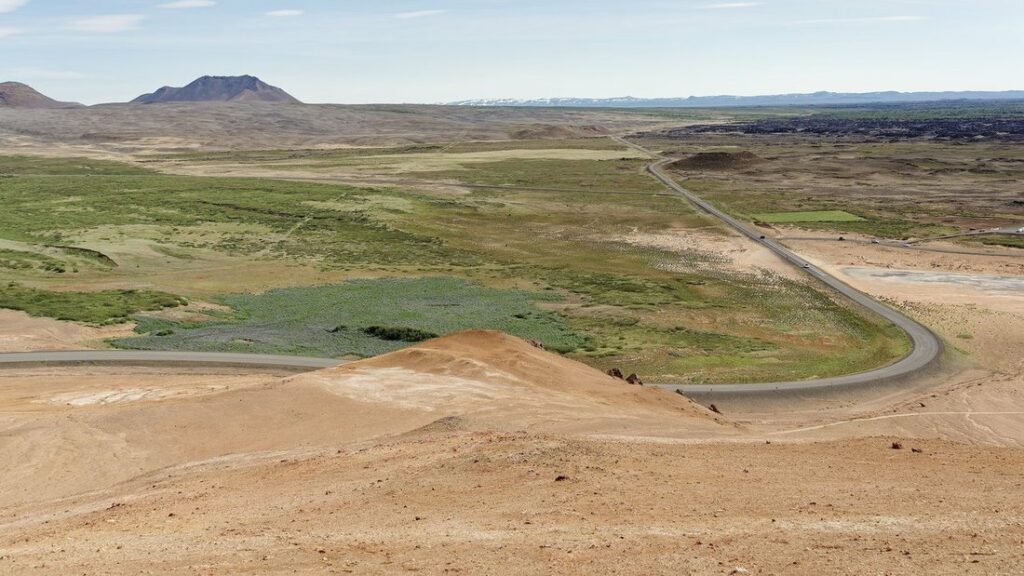 Route asphaltée en courbe traversant une plaine verte et ocre, vue depuis une colline orange, avec un cratère au loin