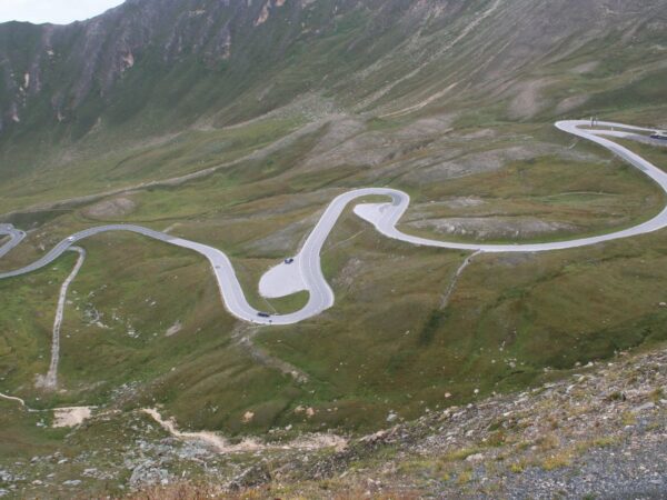 Route de montagne en lacets sur un versant d'herbe rase, avec quelques voitures sur la chaussée
