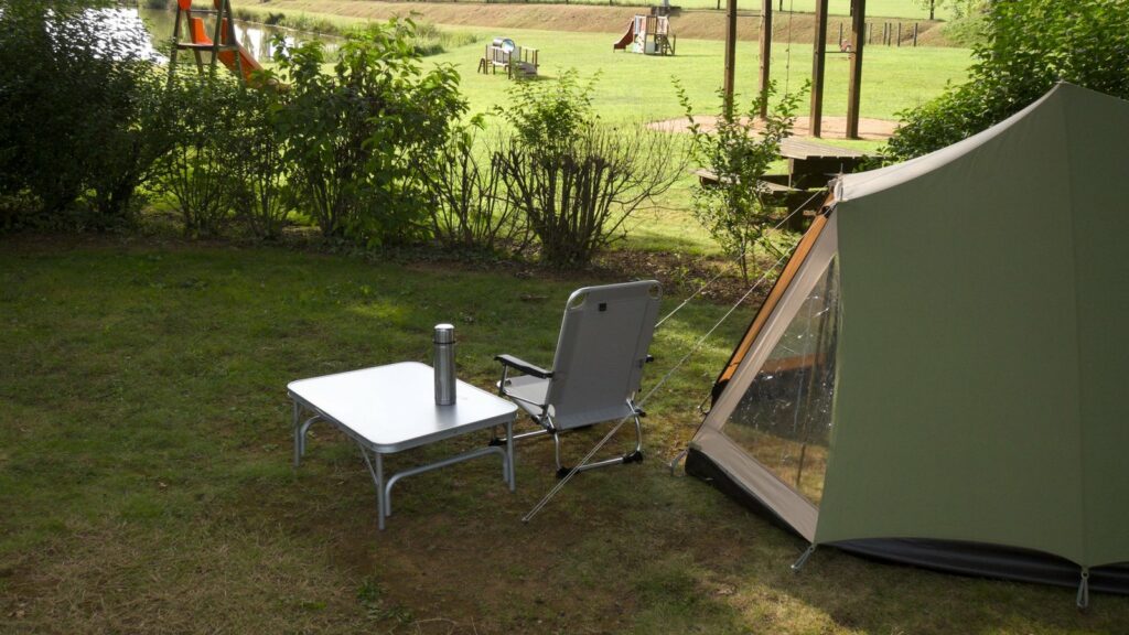Tente verte et beige montée sur une pelouse, avec une table pliante portant un thermos et une chaise pliante, devant une aire de jeux