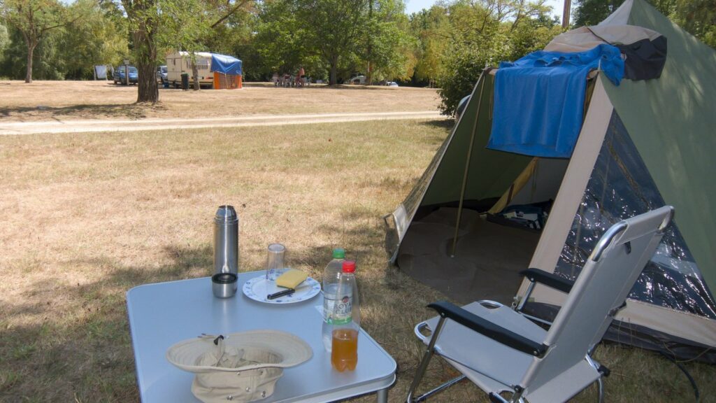 Table pliante avec thermos, assiette et bouteilles, chaise pliante et tente verte sur une pelouse à l'herbe jaunie, caravane au loin