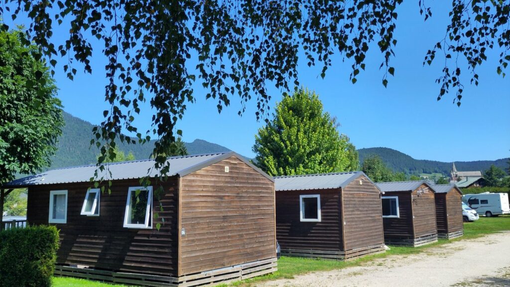 Rangée de chalets locatifs en bois au toit gris sous une branche de bouleau, avec des montagnes boisées derrière