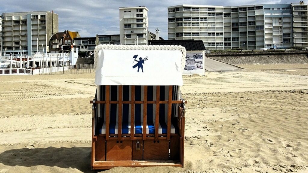 Panier de plage en bois à coussins rayés bleu et blanc, posé sur le sable devant des immeubles du front de mer