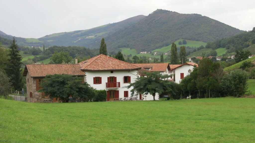 Grande maison blanche aux volets rouges et au toit de tuiles, au bord d'une prairie, avec des collines boisées derrière