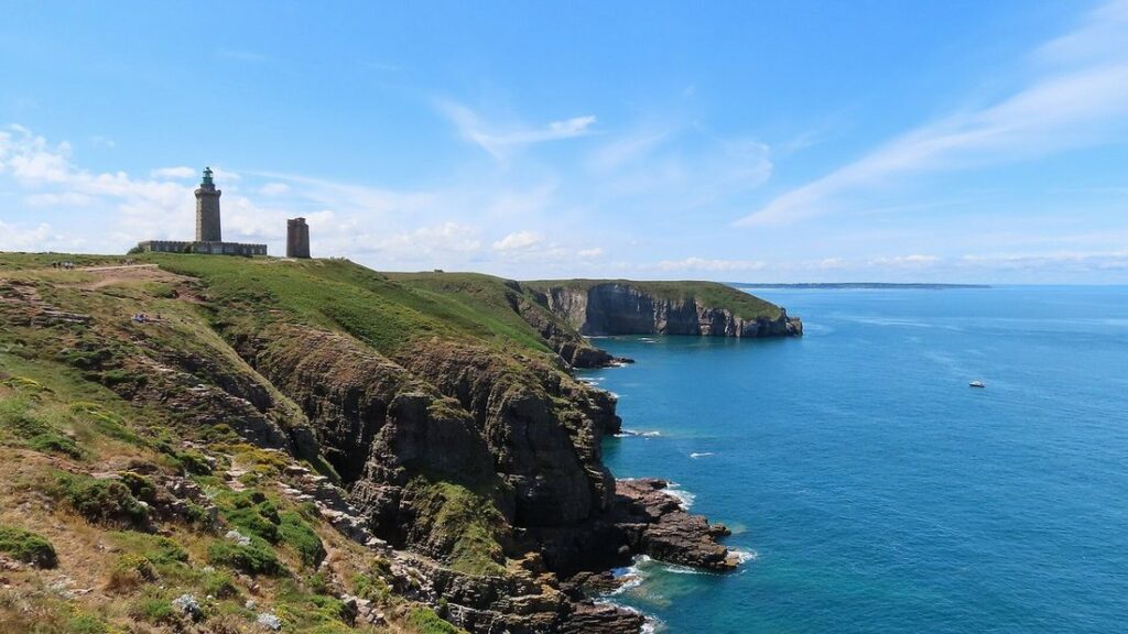 Falaises sombres couvertes de lande dominant une mer bleue, avec un phare et une tour sur le sommet
