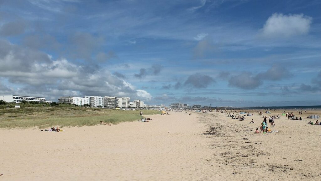 Vaste plage de sable clair longée de dunes herbeuses, avec des immeubles blancs au loin et quelques baigneurs