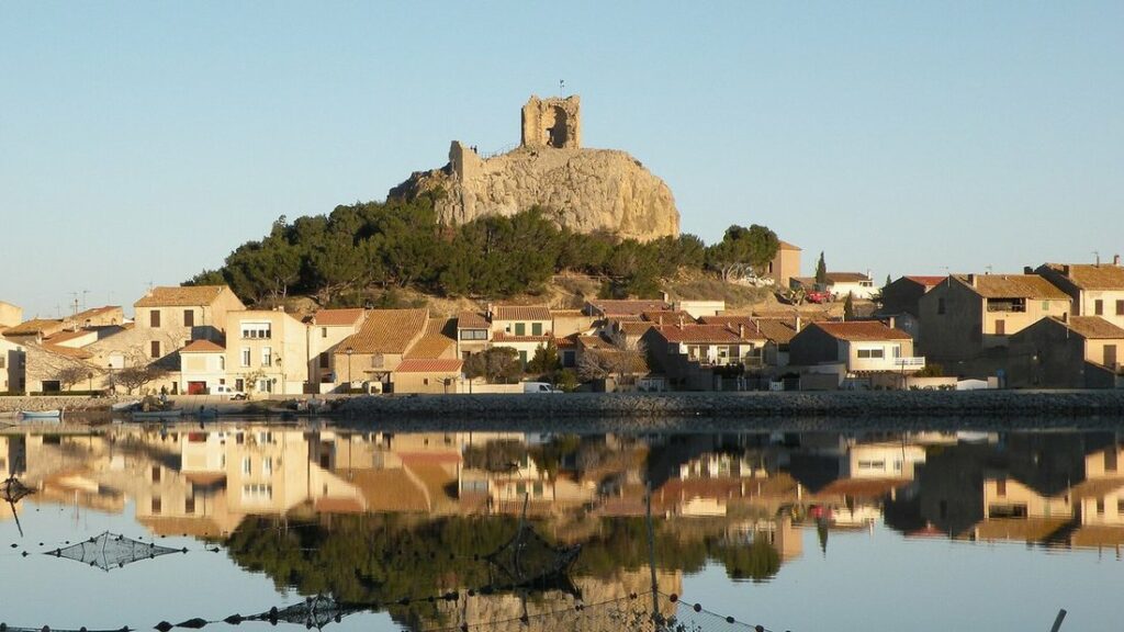 Maisons de village aux toits de tuiles sous un rocher couronné d'une tour en ruine, reflétées dans l'eau d'un étang
