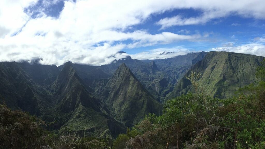 Crêtes vertes aiguës et ravins profonds sous de grands nuages, avec de la végétation au premier plan