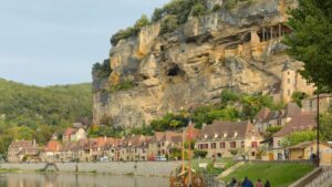 Maisons de pierre dorée alignées au pied d'une falaise, au bord d'une rivière, avec un bateau en bois amarré au premier plan