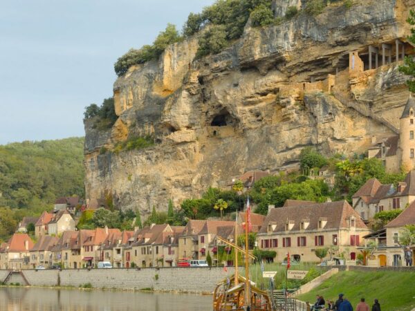 Maisons de pierre dorée alignées au pied d'une falaise, au bord d'une rivière, avec un bateau en bois amarré au premier plan
