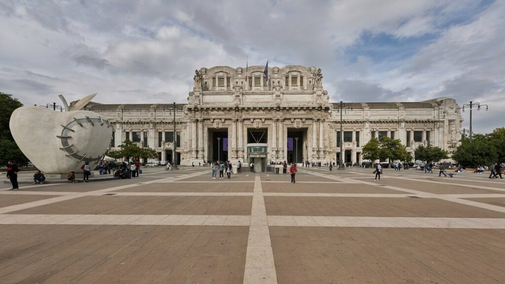 Façade monumentale en pierre claire de la gare Milano Centrale, au bout d'une grande place dallée
