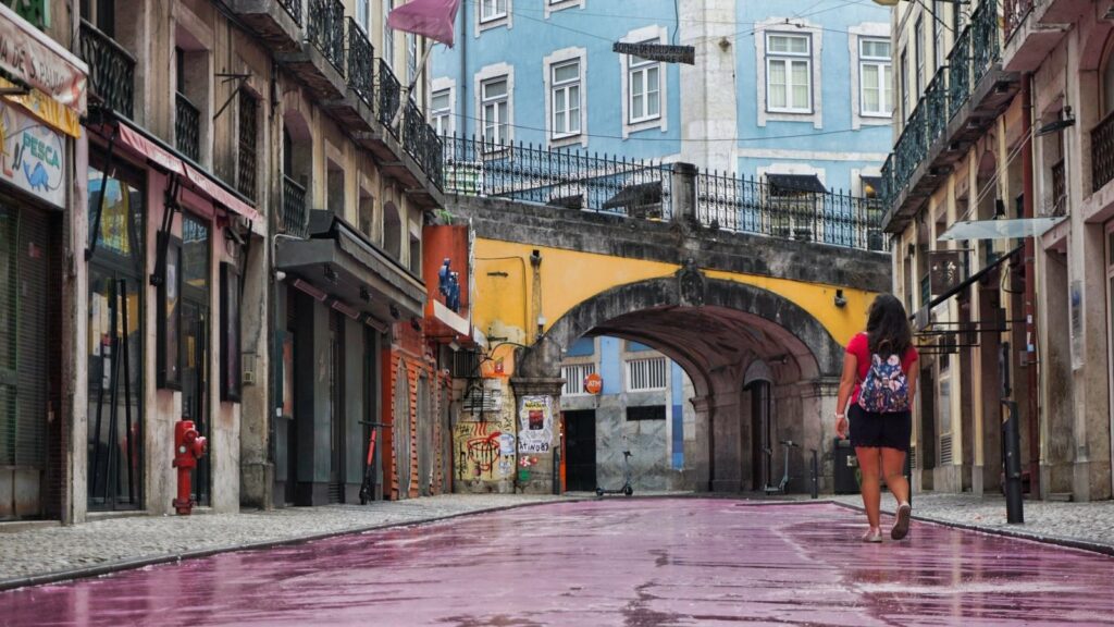 Femme de dos, sac à dos fleuri sur les épaules, marchant dans une rue au pavage rose vers une arche en pierre, entre des façades colorées