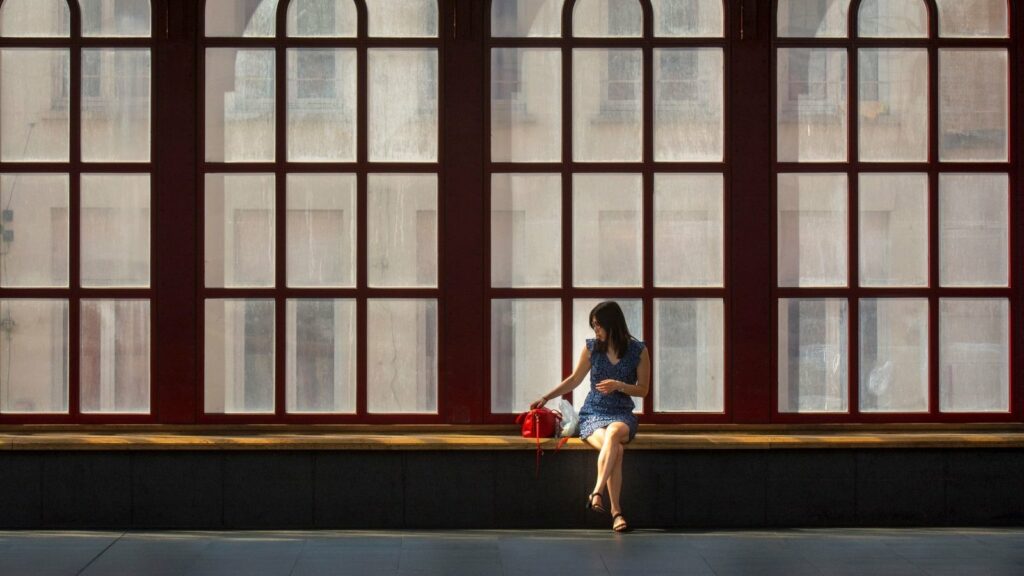 Femme assise seule sur un rebord devant de grandes fenêtres cintrées à petits carreaux, dans un grand hall baigné de lumière