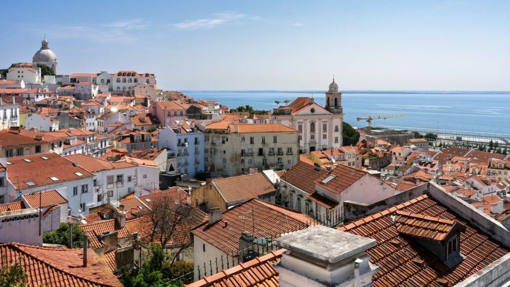 Toits de tuiles orange de l'Alfama à Lisbonne, avec un dôme blanc, un clocher et le Tage au fond